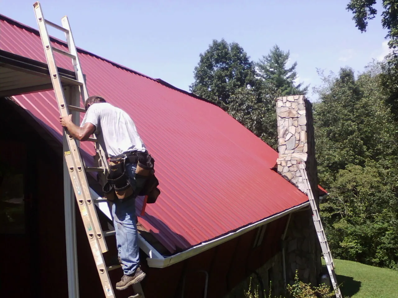 Roofer on a ladder beside a red metal roof and stone chimney — Soffit & Fascia Repair by Roofing Brothers in Laneview